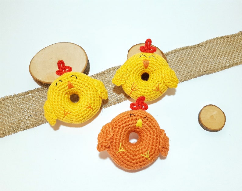 May include: Three crocheted chick toys in the shape of donuts. The chicks are yellow and orange with red beaks and feet.