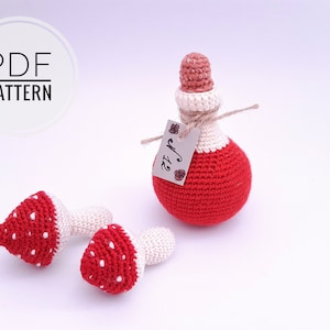 Puede incluir: Botella de poción roja y blanca de ganchillo con un tapón de corcho y una etiqueta que dice "Drink Me". También se muestran dos setas rojas y blancas de ganchillo.