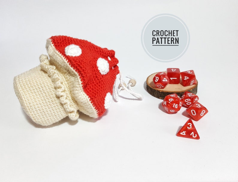 Pode incluir: Saco de dados de croch&ecirc; em forma de cogumelo com fio vermelho e branco, e dados vermelhos com n&uacute;meros brancos. O saco est&aacute; sobre uma superf&iacute;cie branca. CROCHET PATTERN est&aacute; escrito em um c&iacute;rculo cinza.
