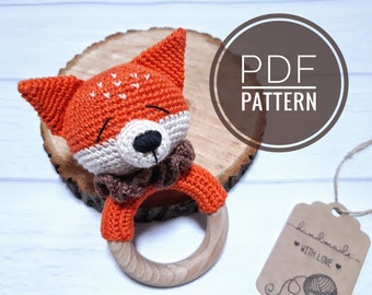 Op de afbeelding: Oranje en witte gehaakte vos-rammelaar met een houten ring. De vos heeft een bruin gehaakt kraagje en zwarte ogen. De rammelaar is handgemaakt en heeft een label met de tekst "handgemaakt met liefde".