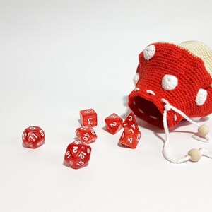 Pode incluir: Um saco de dados de croch&ecirc; em forma de cogumelo vermelho e branco com um cord&atilde;o branco. O saco est&aacute; aberto e dados vermelhos est&atilde;o caindo.