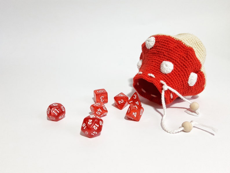 Pode incluir: Um saco de dados de croch&ecirc; em forma de cogumelo vermelho e branco com um cord&atilde;o branco. O saco est&aacute; aberto e dados vermelhos est&atilde;o caindo.
