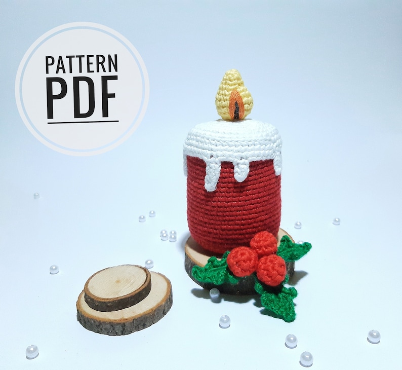 Op de afbeelding: Een gehaakte kaars met een rode body, witte wasdruppels en een gele en oranje vlam. De kaars staat op een houten schijf, versierd met een groene en rode hulsttak. De afbeelding bevat de tekst "PATTERN PDF".