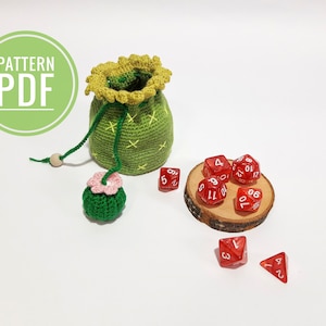 Puede incluir: Una bolsa de dados de crochet verde con forma de cactus con una flor rosa en el lateral. La bolsa está llena de dados rojos con números blancos. La bolsa está sobre una superficie blanca.