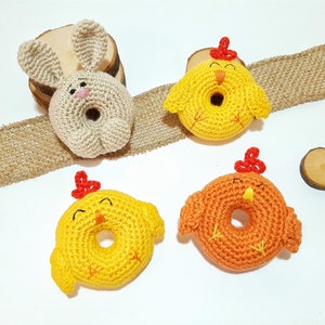 May include: Three crocheted stuffed animals: a beige bunny and two yellow chicks. The bunny has long ears and a small pink nose. The chicks have red combs and beaks. All three animals have a hole in the center, making them look like donuts.
