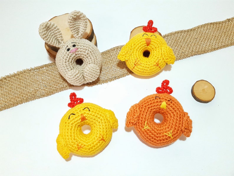 May include: Three crocheted stuffed animals: a beige bunny and two yellow chicks. The bunny has long ears and a small pink nose. The chicks have red combs and beaks. All three animals have a hole in the center, making them look like donuts.