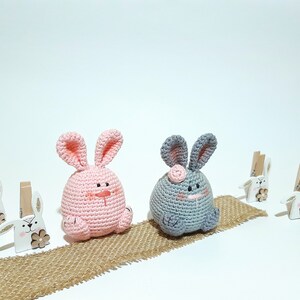 May include: Two crocheted bunny plushies, one pink and one gray, sitting on a burlap surface. The pink bunny has a brown nose and black eyes. The gray bunny has a pink nose and black eyes. Both bunnies have long ears.