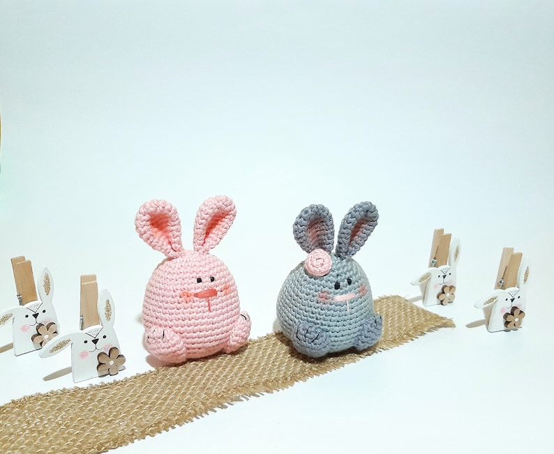 May include: Two crocheted bunny plushies, one pink and one gray, sitting on a burlap surface. The pink bunny has a brown nose and black eyes. The gray bunny has a pink nose and black eyes. Both bunnies have long ears.