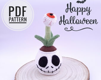 Könnte beinhalten: Eine gehäkelte Halloween-Dekoration mit einem weißen Schädel mit einem gestickten Lächeln und schwarzen Augen. Aus dem Kopf des Schädels wächst eine grüne Pflanze mit einem weißen Augapfel oben drauf. Der Text "Happy Halloween" ist in Schwarz auf weißem Hintergrund geschrieben.
