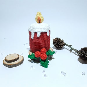 Op de afbeelding: Een handgemaakte gehaakte kaars in rood met een wit wasdruipontwerp. De vlam is geel en oranje. De kaars staat op een houten voet met een gehaakte hulst- en bessenaccent. Een feestelijke kerstversiering.