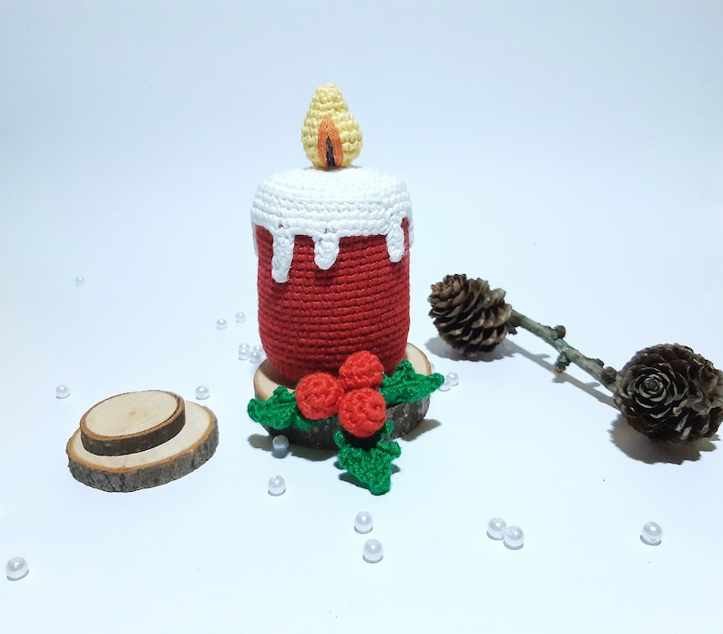 Op de afbeelding: Een handgemaakte gehaakte kaars in rood met een wit wasdruipontwerp. De vlam is geel en oranje. De kaars staat op een houten voet met een gehaakte hulst- en bessenaccent. Een feestelijke kerstversiering.