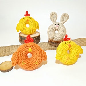 May include: Three crocheted stuffed animals shaped like donuts. One is yellow with a red beak and feet, one is orange with a red beak and feet, and one is beige with long ears and a pink nose.