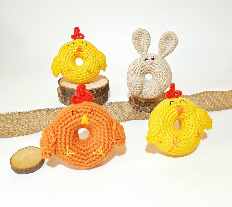 May include: Three crocheted stuffed animals shaped like donuts. One is yellow with a red beak and feet, one is orange with a red beak and feet, and one is beige with long ears and a pink nose.