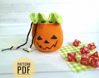 May include: An orange crocheted pumpkin-shaped bag with a green leafy top and black drawstring. The bag has a jack-o'-lantern face and is next to red dice on a green and white checkered cloth. The image includes the text "PATTERN PDF".