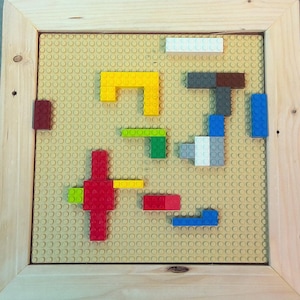 May include: A wooden frame with a beige base plate and colorful Lego blocks arranged in a pattern. The blocks are red, yellow, green, blue, white, and brown.