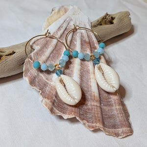 May include: A pair of gold-toned hoop earrings featuring a series of light blue and turquoise faceted beads along the lower curve. Each earring is finished with a single white cowrie shell charm suspended from the centre of the beaded section.