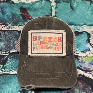 May include: A distressed charcoal gray baseball cap with a mesh back. The front features a rectangular patch with the words "SPEECH LANGUAGE PATHOLOGIST" in colorful letters. The cap has a pre-curved bill and a distressed look.