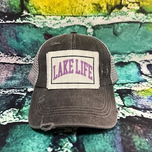 May include: A distressed gray trucker hat with a mesh back and a curved brim. The front patch features the words "LAKE LIFE" in purple letters within a white and black rectangular border. The hat has a worn, vintage look.