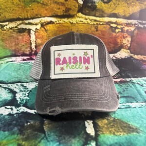 May include: A distressed charcoal gray trucker hat with a white mesh back. The front patch reads "Raisin' Hell" in pink and green lettering with star accents. The hat has a pre-worn look.