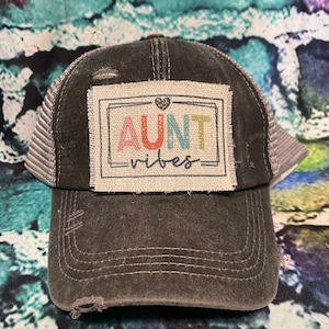May include: A black and gray distressed trucker hat with a patch that says "AUNT vibes" in rainbow colors and a small heart.