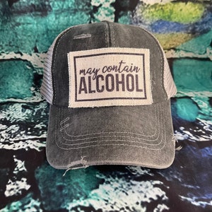 May include: A black and gray distressed trucker hat with a white patch that says "may contain ALCOHOL" in black text.