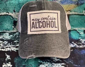 Puede incluir: Una gorra de camionero negra y gris desgastada con un parche blanco que dice "puede contener ALCOHOL" en texto negro.
