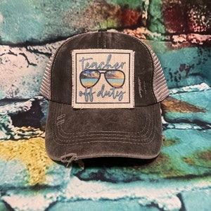 May include: A black distressed trucker hat with a white patch that says "teacher off duty" and features a pair of sunglasses with a beach scene reflected in the lenses.
