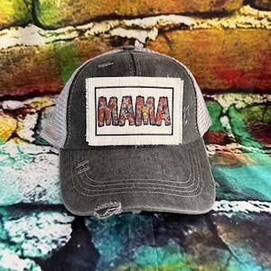 May include: Distressed gray baseball cap with a white mesh back. The front features a rectangular patch with the word "MAMA" in colorful floral letters. The cap has a pre-curved bill and a distressed look.