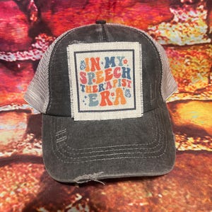 May include: A black and gray distressed trucker hat with a white patch that says "In My Speech Therapist Era" in colorful letters.