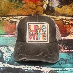 May include: A distressed charcoal gray trucker hat with a mesh back. The front features a square patch with the words "LINE WIFE" in a mix of leopard print and colorful fonts. The hat has a pre-curved brim and a worn appearance.