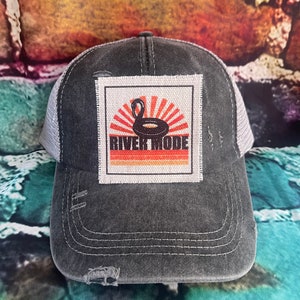 May include: A black and gray distressed trucker hat with a white patch featuring a black inner tube with a swan on top, a red and orange sunburst design, and the text "River Mode".