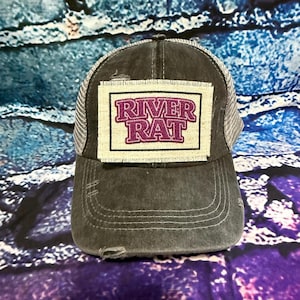 May include: A distressed gray trucker hat with a mesh back and a curved brim. The hat features a rectangular patch with the words "RIVER RAT" in purple block letters. The hat has a worn, vintage look.