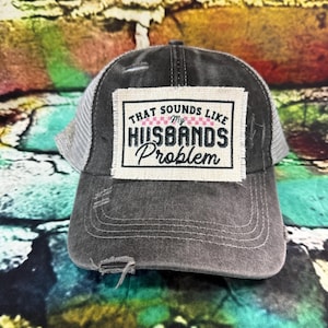 May include: A distressed gray trucker hat with a white mesh back. The front patch reads "THAT SOUNDS LIKE MY HUSBANDS PROBLEM" in black and pink lettering. The hat has a pre-curved brim and a distressed look.