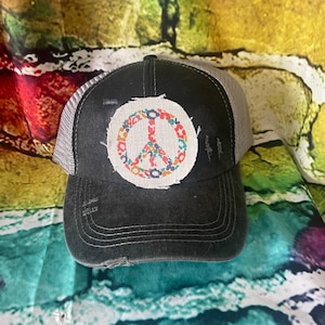 May include: A black trucker hat with a distressed look. The hat features a circular patch with a peace sign design in a variety of colors. The mesh back is gray.