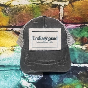 May include: A black and gray distressed trucker hat with a beige patch that reads "Undiagnosed but somethin' ain't right".