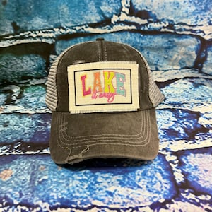 May include: A charcoal gray distressed baseball cap with a mesh back and a rectangular patch on the front. The patch features the text "LAKE it easy" in colorful, multi-toned block lettering with a frayed fabric edge and black border.