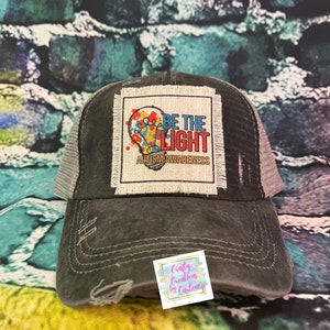 May include: A black distressed trucker hat with a white patch that says "Be the Light Autism Awareness" with a colorful lightbulb graphic.