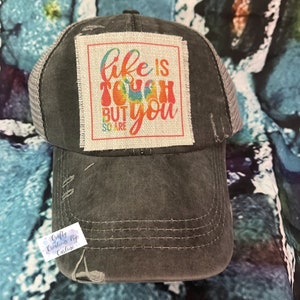May include: A black and gray distressed trucker hat with a square patch that reads "Life is tough but you so are". The patch has a rainbow tie-dye design.