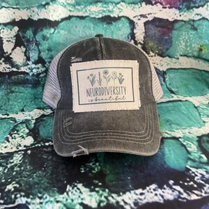 May include: A black and gray distressed trucker hat with a white patch that says "Neurodiversity is beautiful" with a small floral design.