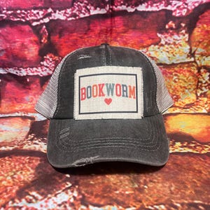 May include: A black and gray distressed trucker hat with a white patch that says "BOOKWORM" in red, blue, and white letters with a red heart.