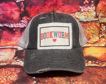 Könnte beinhalten: Eine schwarze und graue, abgenutzte Trucker-Kappe mit einem weißen Patch, auf dem "BOOKWORM" in roten, blauen und weißen Buchstaben mit einem roten Herzen steht.