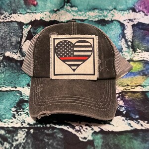 May include: Distressed charcoal gray trucker hat with a white mesh back. The front features a square patch with a heart-shaped American flag design and a red stripe. The hat has a pre-worn look with distressed details.