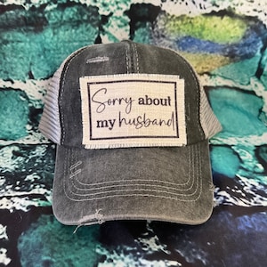 May include: A black and gray distressed trucker hat with a white patch that says "Sorry about my husband".
