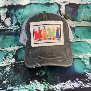 May include: A black and white trucker hat with a distressed look. The hat has a patch on the front that says "AUTISM MAMA" in rainbow colors.