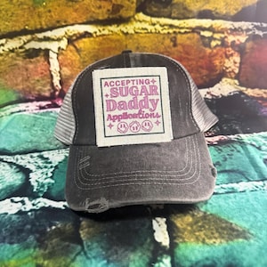 May include: A black and gray distressed trucker hat with a white patch that says "Accepting Sugar Daddy Applications" in pink lettering with three pink smiley faces.