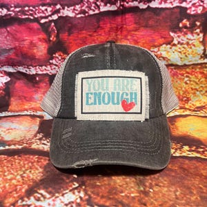 May include: A black and gray distressed trucker hat with a patch that says "You Are Enough" in teal lettering with a red heart.