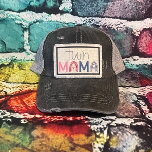May include: A black and gray distressed trucker hat with a patch that says "Twin Mama" in colorful letters with a rainbow design.