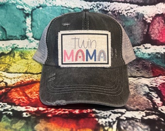 May include: A black and gray distressed trucker hat with a patch that says "Twin Mama" in colorful letters with a rainbow design.