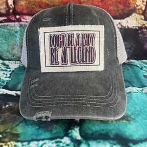 May include: A black and gray trucker hat with a white patch that says "Don't be a lady be a legend" in pink and black lettering.