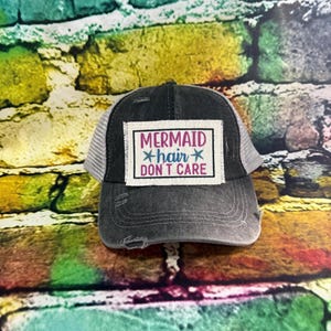 May include: A distressed black trucker hat with a white mesh back. The front patch reads "MERMAID hair DON'T CARE" in pink and blue text. The hat has a curved brim and a faded, worn look.